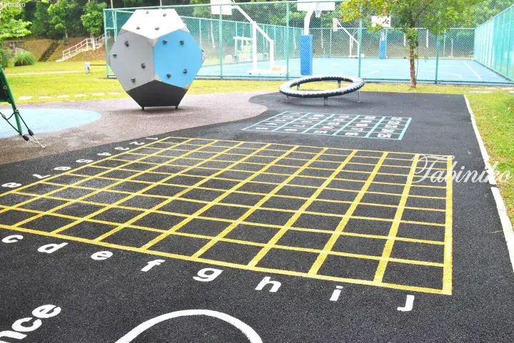 Children’s playground features a large yellow grid game painted on black rubber flooring, alongside a climbing structure and trampoline.