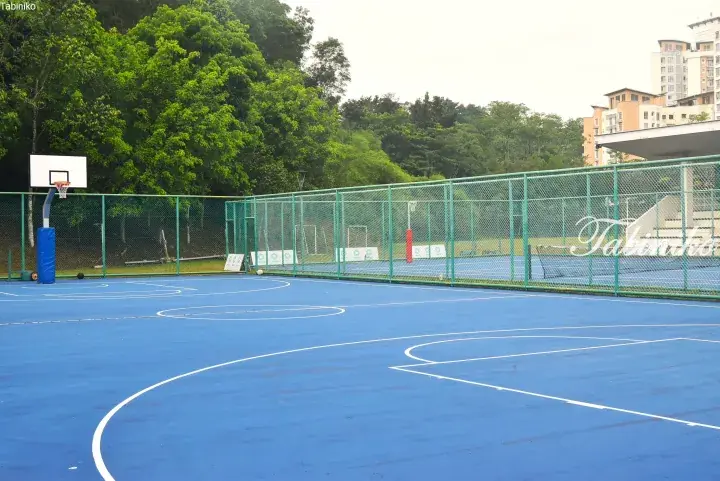 Blue outdoor basketball court with green fencing, surrounded by lush trees and residential buildings.