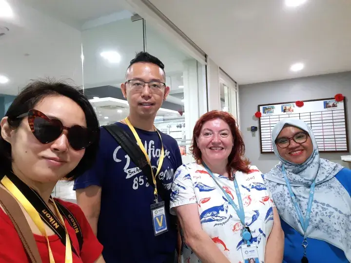Four diverse travelers wearing visitor badges and lanyards pose for a selfie in a modern office corridor.
