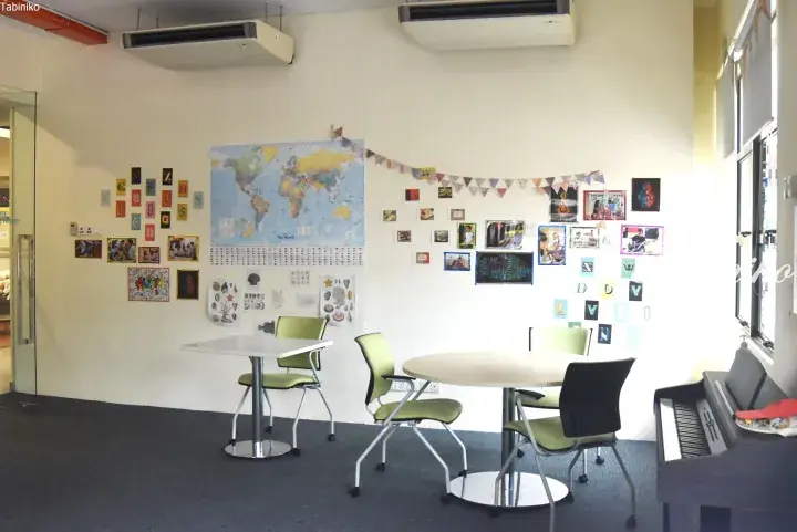 Bright classroom with world map, student photos, and green chairs around tables near a piano.