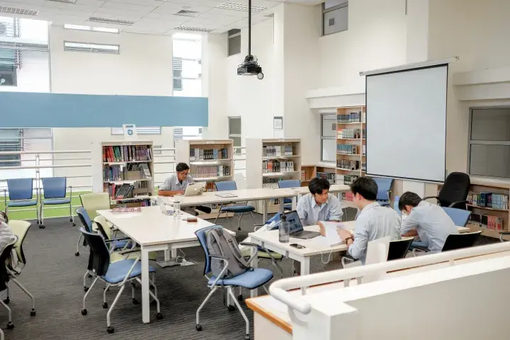 Students studying with laptops and books in a bright university library with bookshelves and a projector screen.