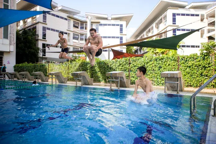 Children joyfully jumping into a resort swimming pool with starting blocks and modern apartment buildings in the background.