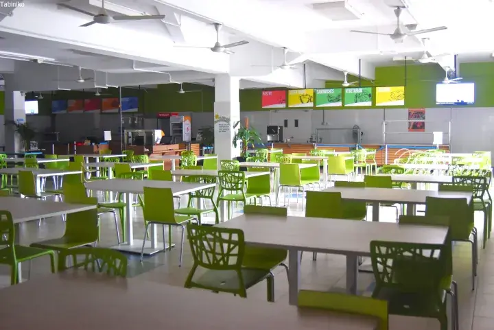 Bright university cafeteria with lime green chairs and white tables, featuring food stall signs for pastries and sandwiches.