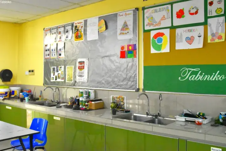 Bright classroom with yellow walls, green cabinets, sinks, and student art including Malaysia Merdeka drawings on bulletin boards.