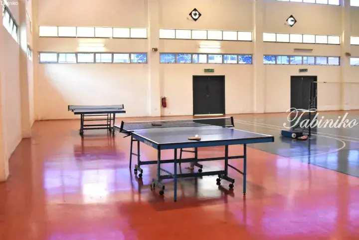 Indoor sports hall with red flooring featuring two blue table tennis tables and a badminton court area.