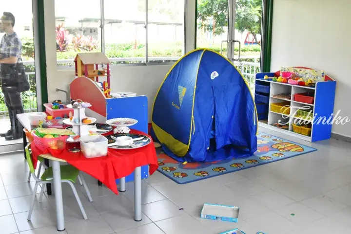 Colorful children’s play area featuring a blue tent, toy kitchen, and table set with play food in a bright room.