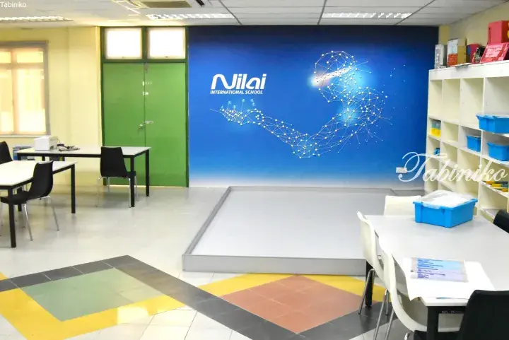 Nilai International School classroom featuring a blue wall with a geometric art design, tables, chairs, and storage shelves.