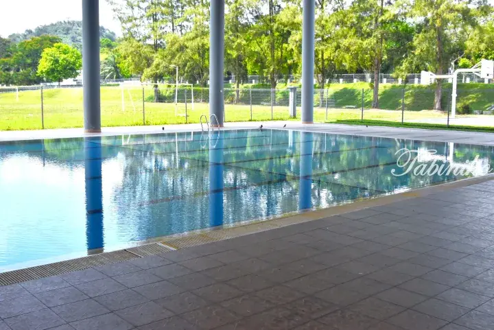 Covered outdoor swimming pool with blue pillars and lane markings, surrounded by a green sports field and trees.