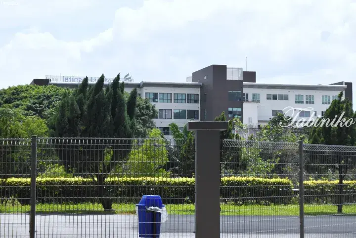 Modern white and brown building behind a metal fence with lush green trees and a blue trash bin in the foreground.