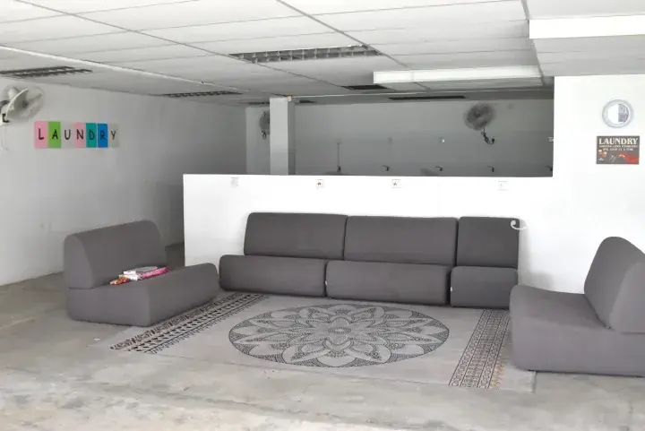 Grey modular sofa set arranged on a patterned rug in a laundry facility with wall-mounted fans and colorful signage.