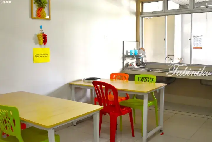 Bright communal pantry with yellow tables, colorful chairs, and a sink area in a Malaysia student hostel.