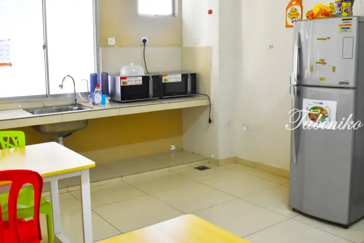 Shared kitchen area with microwave, refrigerator, sink, and colorful chairs in a Malaysian rental property.