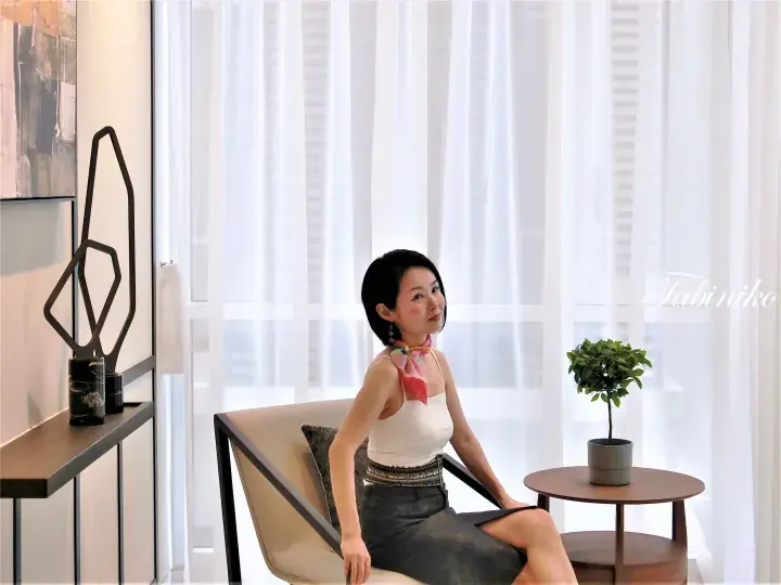 Woman in white top and dark skirt sitting in modern chair beside sheer curtains in a stylish property interior.