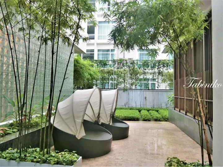 Private balcony with two cushioned daybeds and bamboo plants overlooking a modern hotel building.