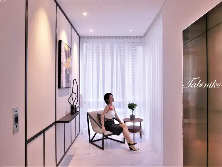 Woman in white top and black skirt sitting on a modern chair in a bright, minimalist hallway with sheer curtains.