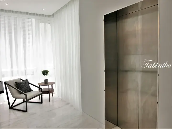 Modern minimalist interior with a sleek lounge chair, small table, and floor-to-ceiling white curtains beside a metallic elevator door.