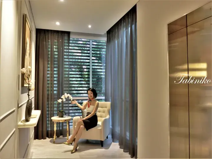 Woman in a white top and black skirt sitting on a cream armchair in a modern hotel room with large windows and gold accents.