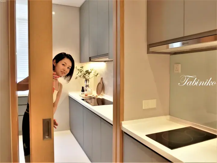 Woman peeking from behind a wooden door into a modern kitchen with grey cabinets and an Electrolux range hood.