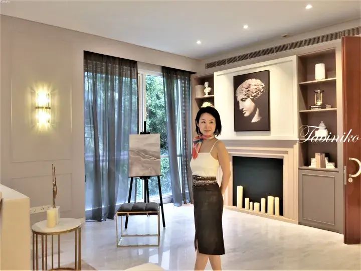 Woman in white top and black skirt standing in a modern luxury living room with marble floors, fireplace, and large windows.