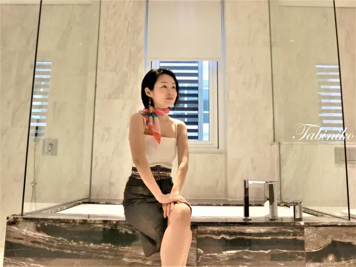 Woman sitting on marble bathtub edge in luxury bathroom with glass shower and window blinds