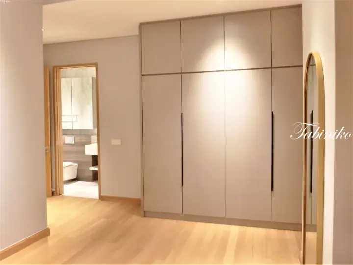 Modern built-in wardrobe with sleek handles in a minimalist bedroom with light wood flooring and an en-suite bathroom.