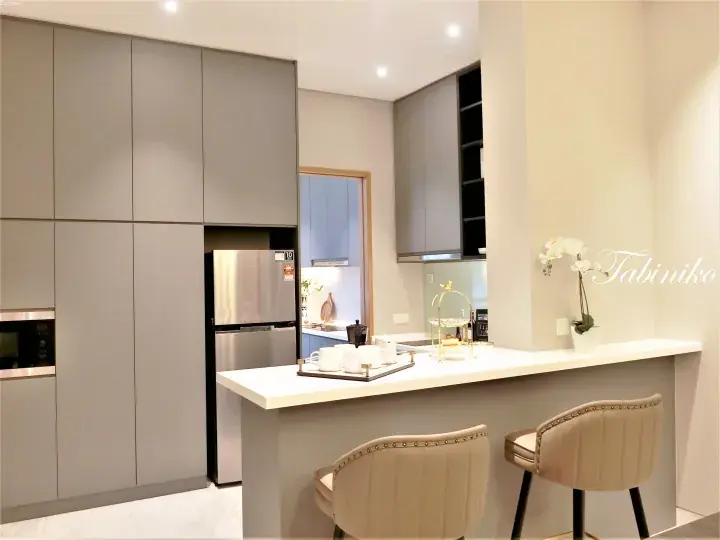 Modern kitchen with grey cabinets, stainless steel fridge, and white breakfast bar with two beige chairs.