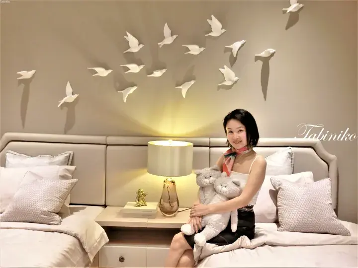 Woman holding stuffed cats on a bed with a lamp and white bird wall art in a modern bedroom.