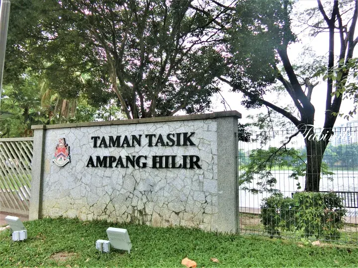 Entrance sign for Taman Tasik Ampang Hilir park featuring the Kuala Lumpur city crest against a backdrop of lush greenery and a lake.