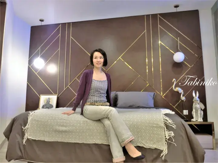 Woman sitting on a bed in a modern bedroom with a geometric gold-accented headboard wall and hanging pendant lights.
