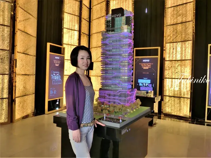Woman standing beside a lit architectural model of the NOVO high-rise building at a property showroom.
