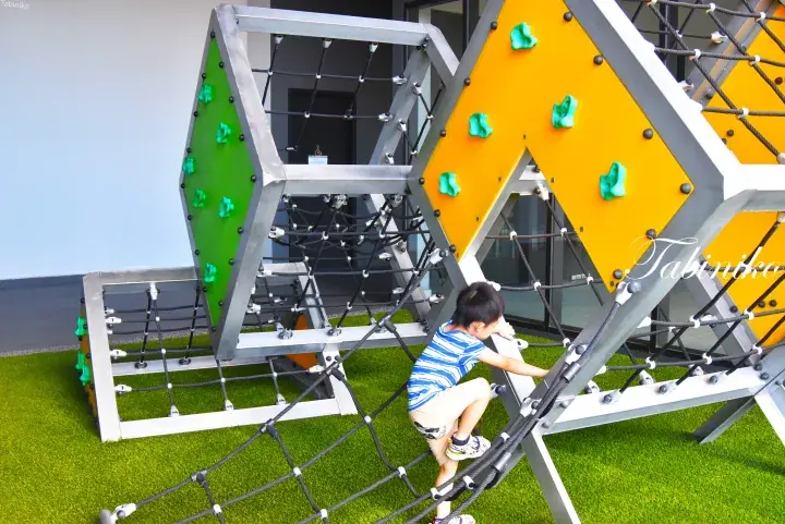 人工芝の上で、ロープネットや緑色のホールドが付いたカラフルな屋外遊具に登る男の子。