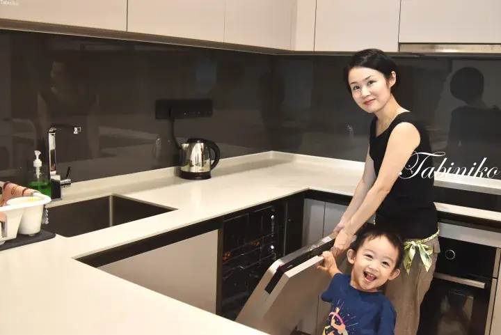 白いカウンターとダークな壁紙が特徴的なモダンなキッチンで、女性と幼い子供が食洗機を開けて笑顔を見せる。