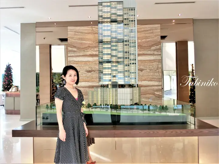 Woman in polka-dot dress stands beside a detailed architectural model of a modern high-rise building in a hotel lobby.
