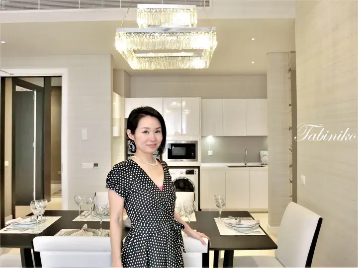 Woman in polka-dot dress standing in a modern dining area with a crystal chandelier and kitchen.