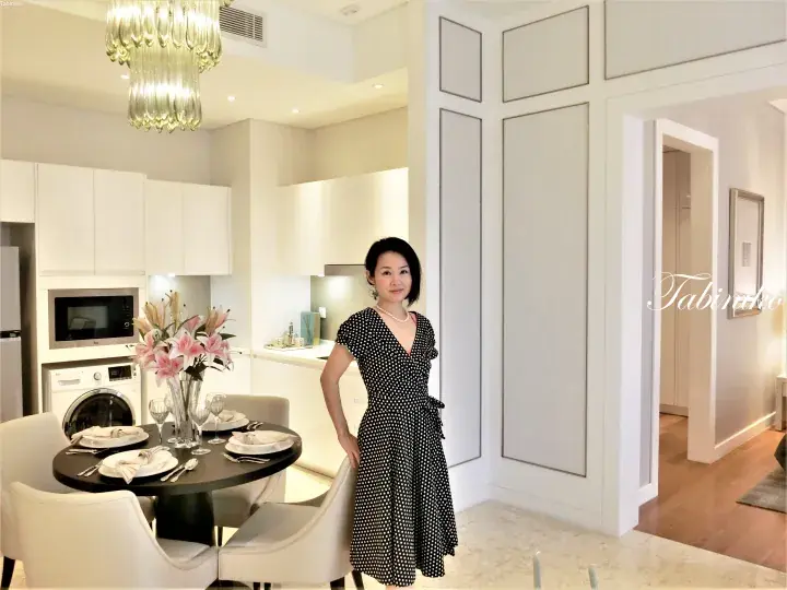 Woman in polka dot dress standing in a modern white kitchen with dining table and chandelier.
