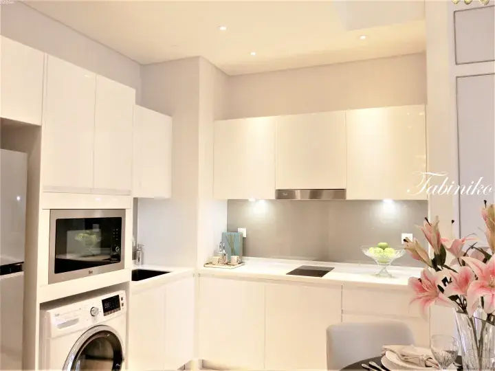 Modern white kitchen with built-in microwave and washing machine, induction cooktop, and pink lilies on dining table