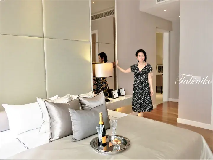 Woman in polka-dot dress standing beside a bed with champagne and chocolates in a luxury hotel room.