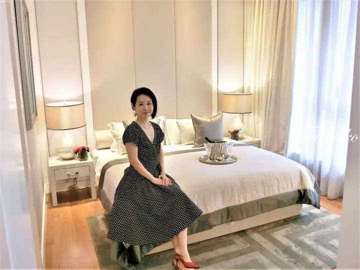 Woman in polka dot dress sitting on a hotel bed with champagne tray in a bright room.