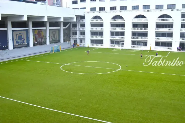 Students playing football on a green artificial turf field surrounded by a white multi-story school building.