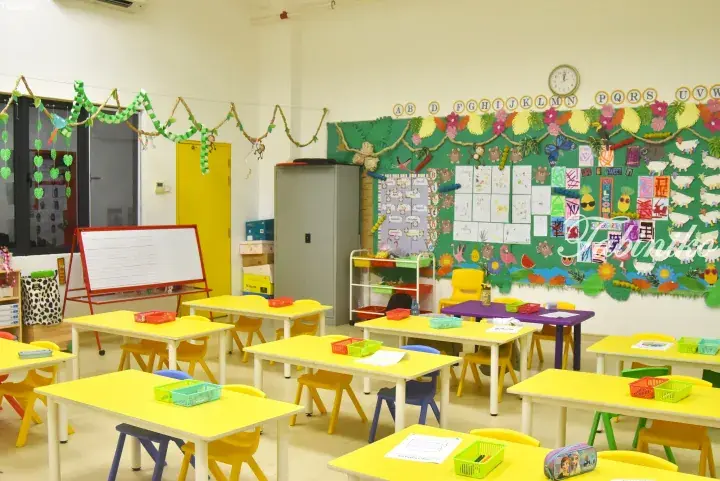 Bright kindergarten classroom with yellow tables, colorful chairs, and a jungle-themed bulletin board displaying children’s artwork.
