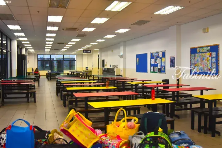 Colorful school cafeteria with rows of yellow and red tables, benches, and backpacks in the foreground.