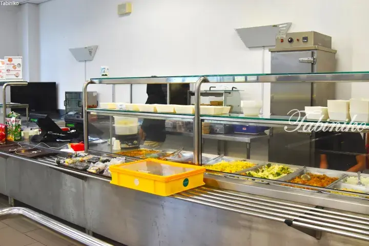 Food counter with stainless steel trays of yellow rice, vegetables, and curry in a cafeteria setting.