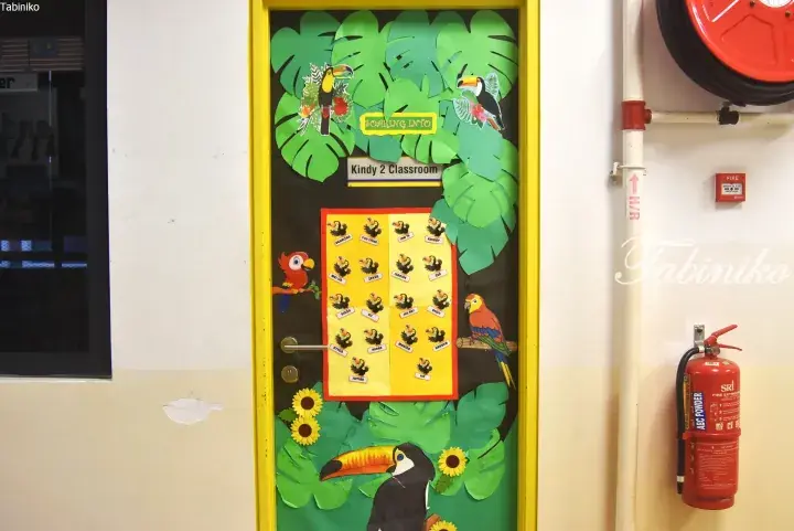 Kindy 2 classroom door decorated with toucans and leaves, featuring a student name chart and a fire extinguisher nearby.