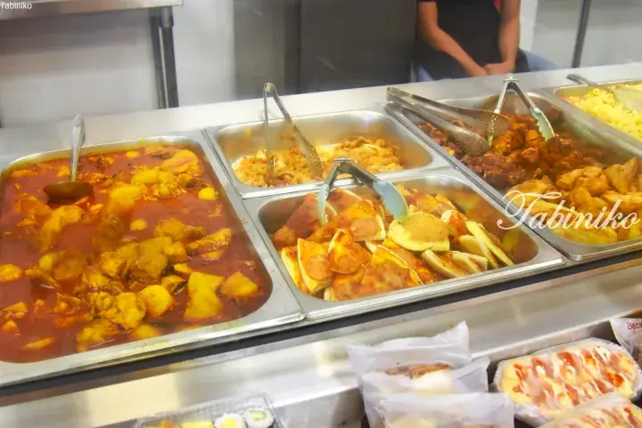 Buffet display with trays of curry chicken, pizza slices, fried chicken, and pasta at a Malaysian eatery.