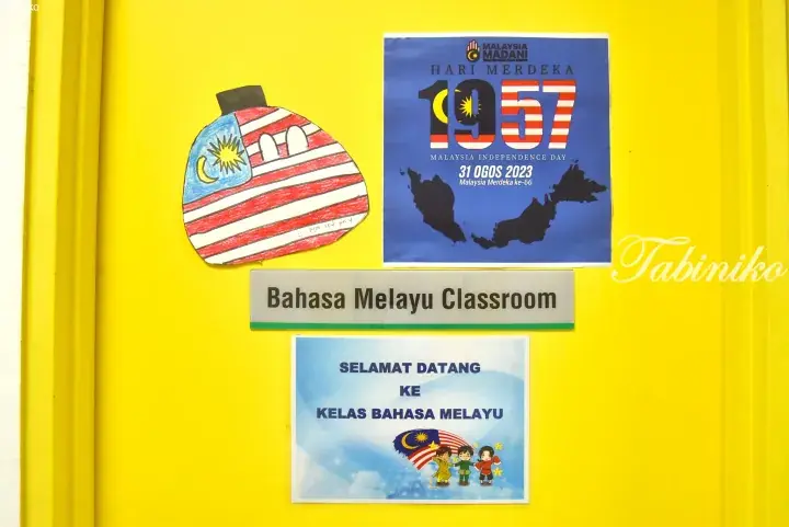 Yellow door of a Bahasa Melayu classroom decorated with Merdeka 2023 posters and a student’s flag artwork.