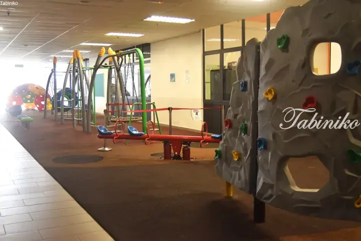 Indoor playground featuring a climbing wall, seesaw, and swings in a modern facility.