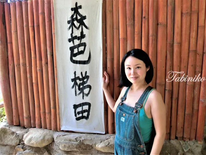 Woman standing beside a bamboo wall with a sign reading ‘Sen Bata Terrace’ at a Malaysian rural retreat.