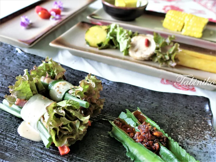 Assorted fresh spring rolls and vegetable wraps served on a dark rectangular plate with corn and salad in the background.