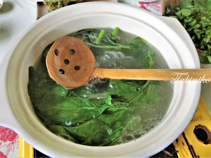 Wooden ladle stirring fresh green vegetables in a white ceramic pot on a portable stove.