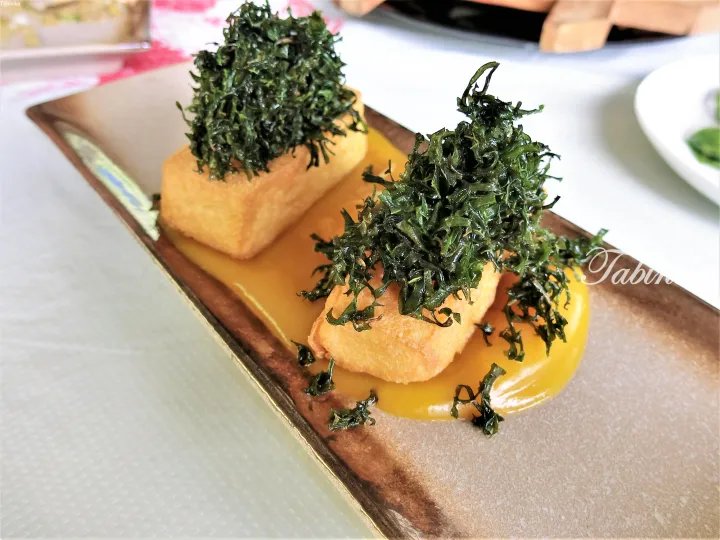Fried tofu topped with crispy green herbs served on a yellow sauce in a rectangular ceramic dish.